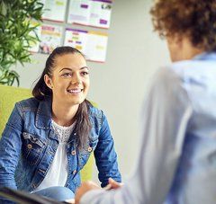 jeune fille souriante et déontractée assise sur un fauteuil parle avec une personne de dos dans un lieu d'accueil