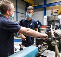 Un homme masqué discute avec son jeune apprenti masqué à côté de machines industrielles